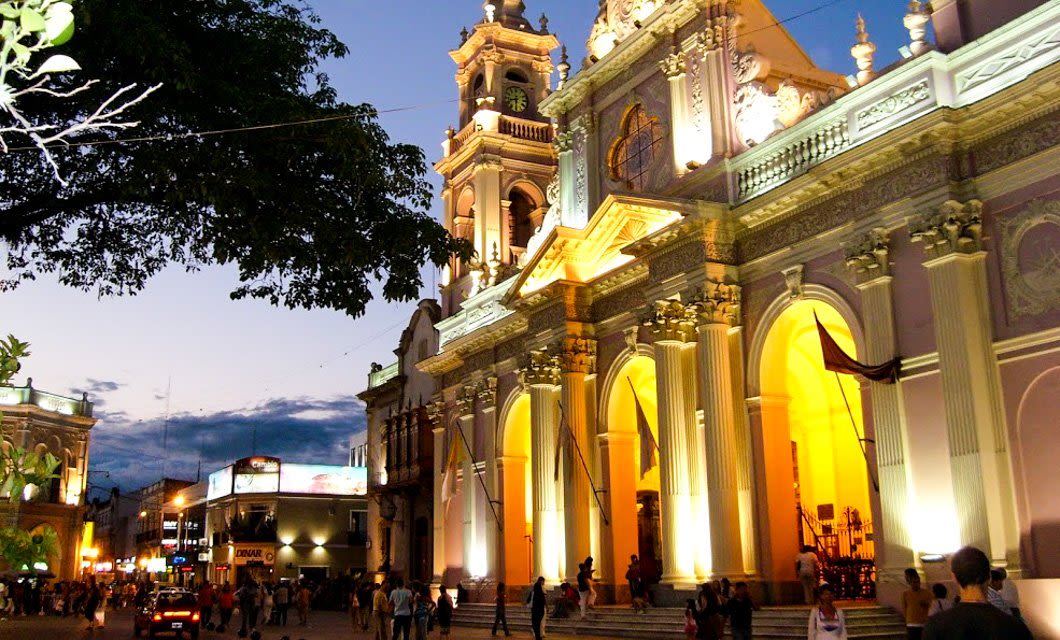 A brightly lit colonial-style cathedral with ornate arches and a tall bell tower stands in Cafayate’s lively city square at dusk, along Salta’s famous Wine Route, as people stroll beneath a sky turning dark blue.