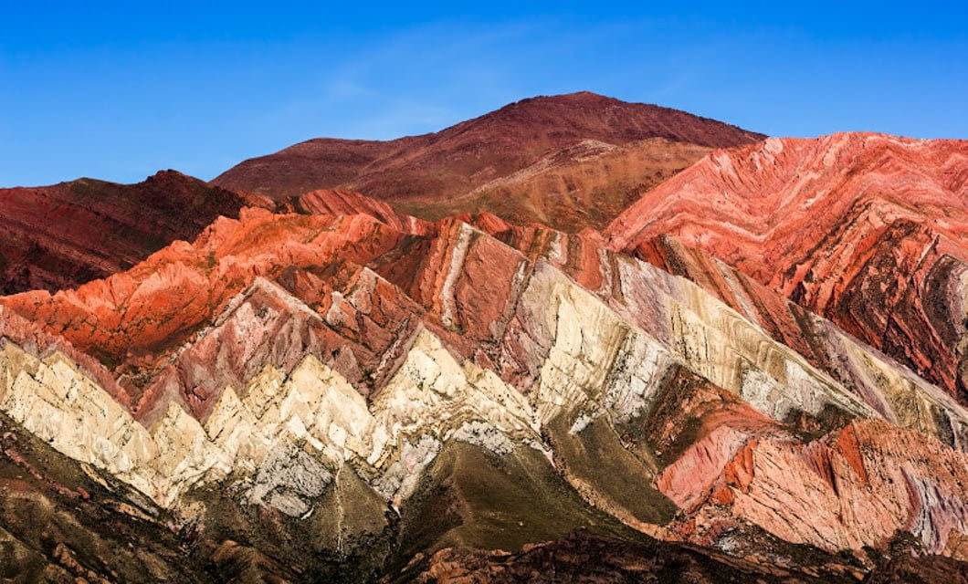 Colorful mountain range with dramatic, jagged stripes of red, pink, orange, yellow, and white rock layers beneath a clear blue sky—an iconic highlight on a Coast to Coast Tour. The landscape shows bold, slanted geological formations.