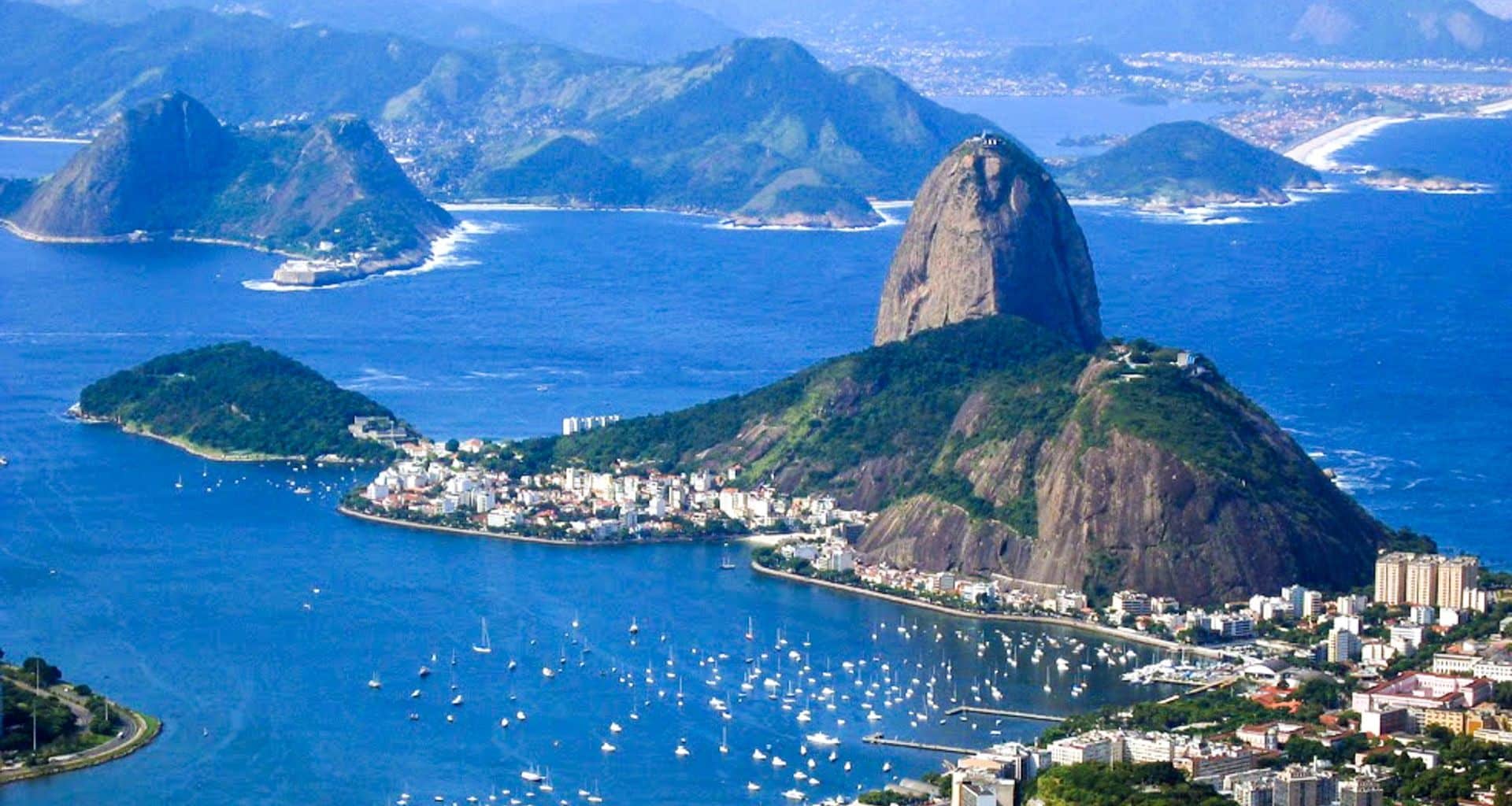 Aerial view of Rio de Janeiro’s Sugarloaf Mountain, surrounded by water and boats, with the city, green hills, distant beaches and mountains in the background—capturing the vibrant spirit of samba in this breathtaking scene.