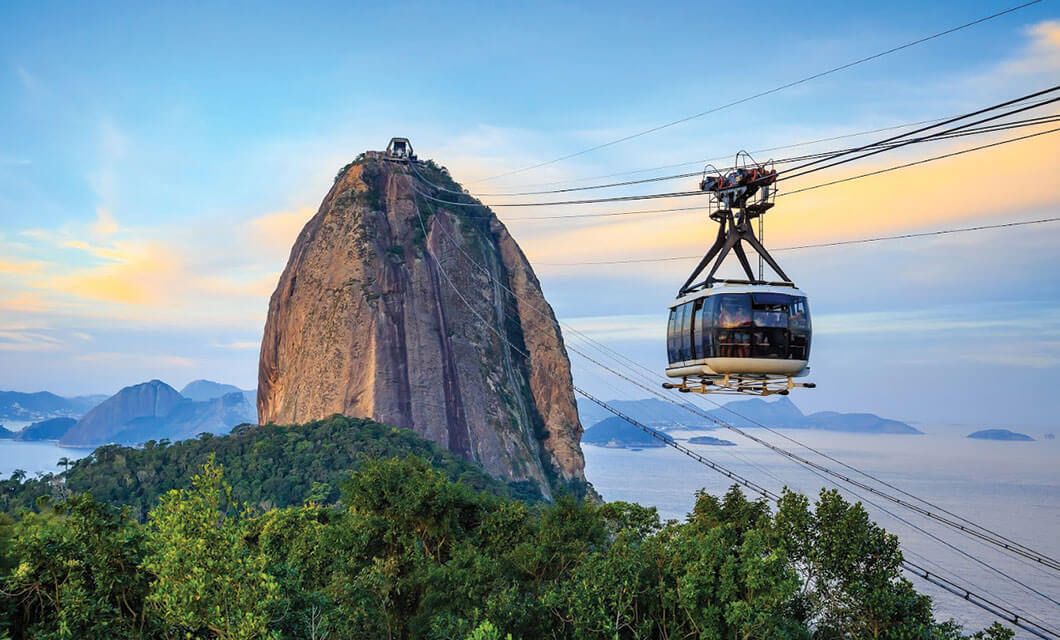 A cable car travels across wires toward Sugarloaf Mountain in Rio de Janeiro, Brazil, with lush greenery below and the ocean and distant hills visible under a colorful sky at sunset—a perfect moment on a Brazil Tour adventure.
