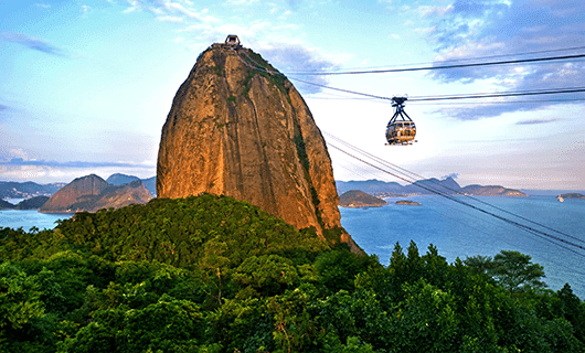 A cable car travels to the top of Sugarloaf Mountain, rising above lush greenery and overlooking the ocean and distant mountains on a scenic tour under a partly cloudy sky in Rio de Janeiro, Brazil.