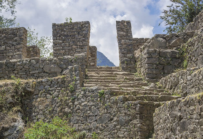 Stone steps lead up through ancient stone walls and doorways at a South America ruin site, with greenery growing around the structures—a perfect spot for trekking—with a mountain visible in the background under a partly cloudy sky.