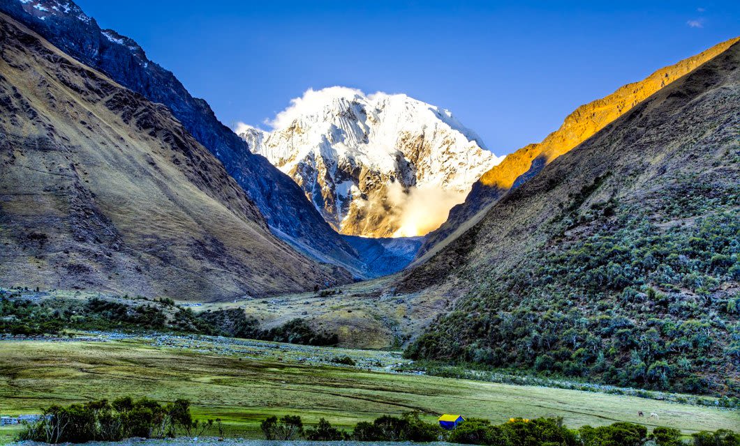 Snow-capped mountain peak framed by steep, sunlit valley slopes, with a lush green meadow and a small tent—an inviting scene along the Salkantay Trek to Machu Picchu under a clear blue sky.