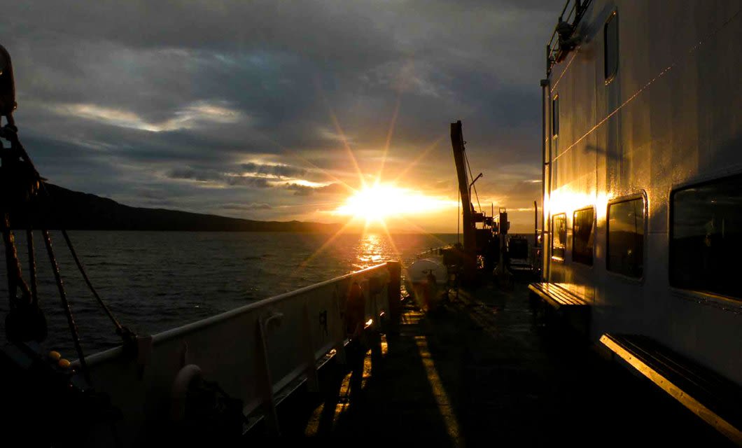 Sunset view from a ship’s deck during an Antarctica Ushuaia cruise, with the sun low over the horizon casting golden reflections on the water and ship’s side, framed by dark clouds overhead.