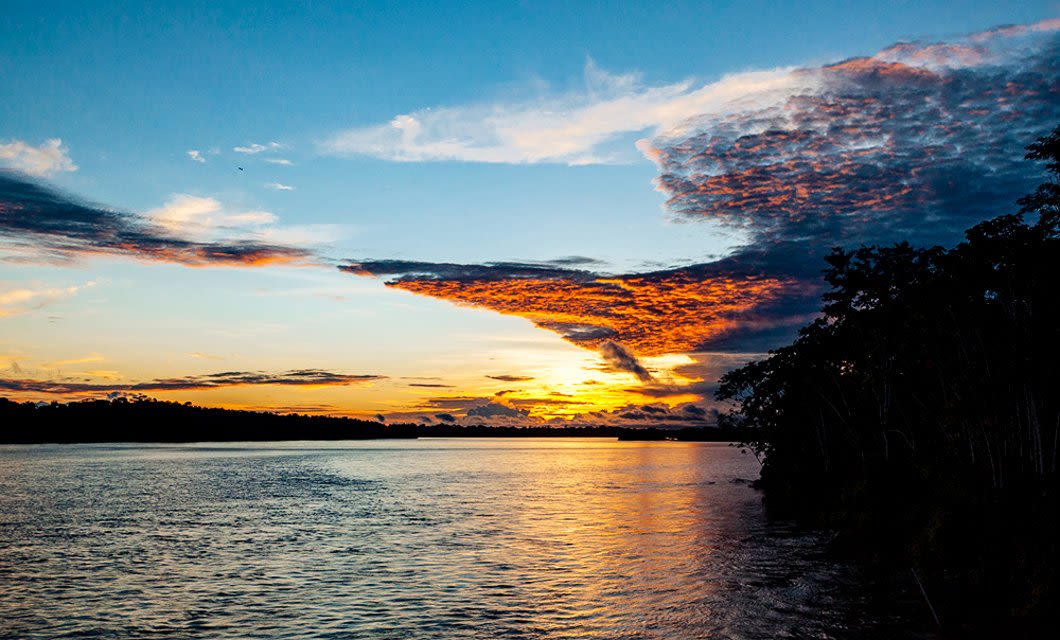 Sunset over the wide Amazon River with vibrant orange and blue clouds reflecting on the water. Silhouettes of trees line the riverbank on the right, capturing the serene beauty often experienced on Amazon Cruises.