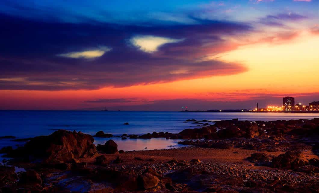 Rocky shoreline at dusk with a colorful sunset sky, blue and orange hues, calm sea, and distant city lights illuminating buildings along the horizon—capturing the tranquil beauty of Uruguay.