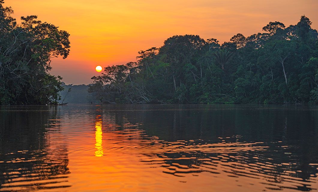 The sun sets over the calm Amazon River, surrounded by dense, green rainforest. The orange and yellow sky reflects on the water’s surface, creating a serene atmosphere—an unforgettable scene on a Brazil Amazon River Cruise.