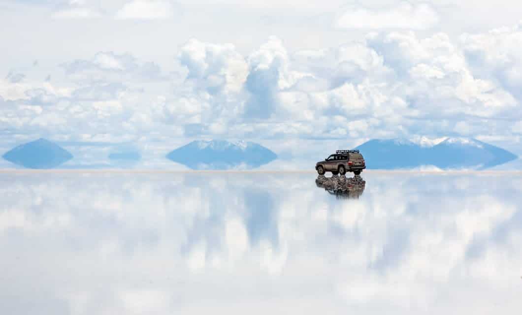 A solitary SUV is parked on a vast, reflective salt flat beneath a cloudy Andes sky, with distant mountains mirrored in the surface, creating a surreal, dreamlike landscape.