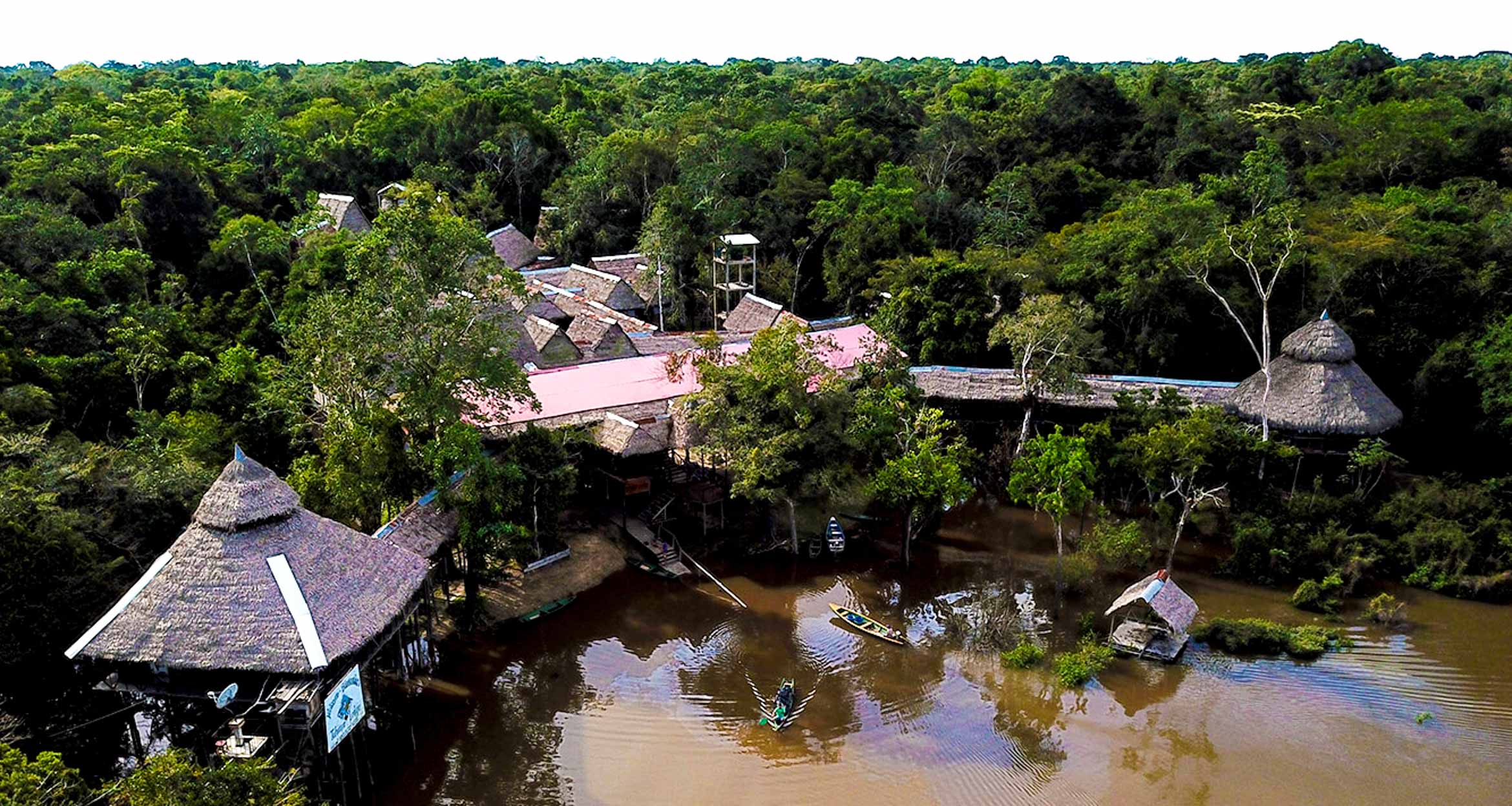 Aerial view of Tahuayo Lodge with thatched roofs nestled in dense rainforest, alongside a muddy river where small boats for Amazon expeditions and wildlife tours are docked near the shore.