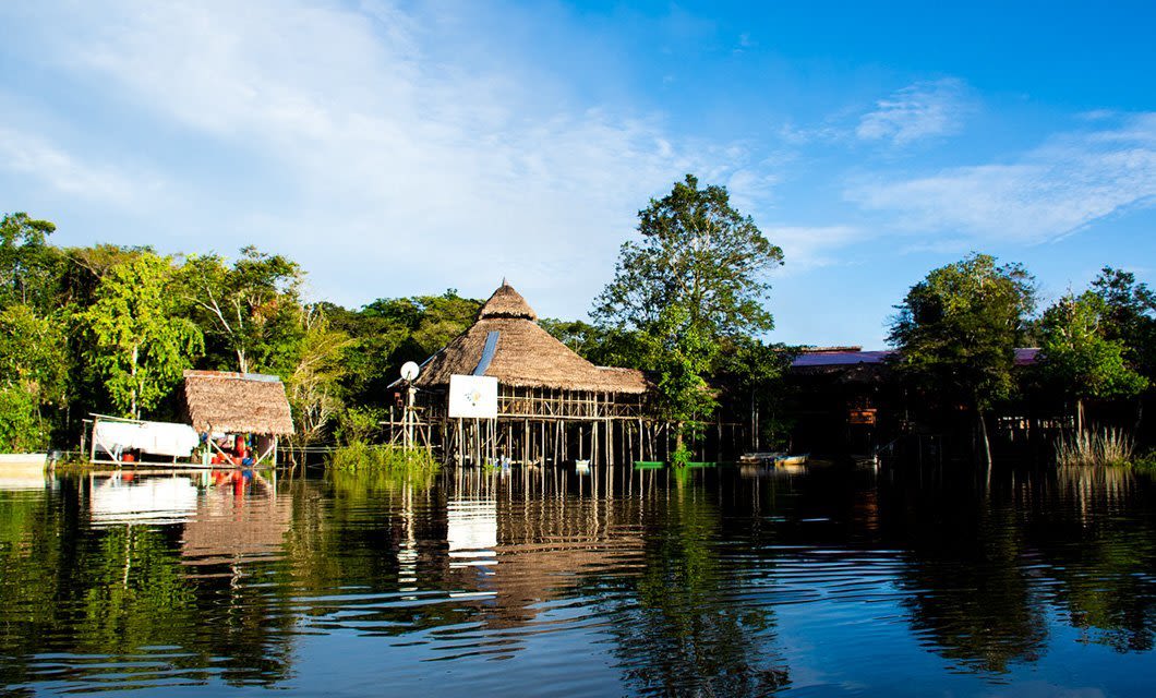 A thatched-roof building on stilts, reminiscent of Tahuayo Lodge, stands by a calm river surrounded by lush green trees under a bright blue sky—a serene setting perfect for Amazon expeditions and wildlife tours.