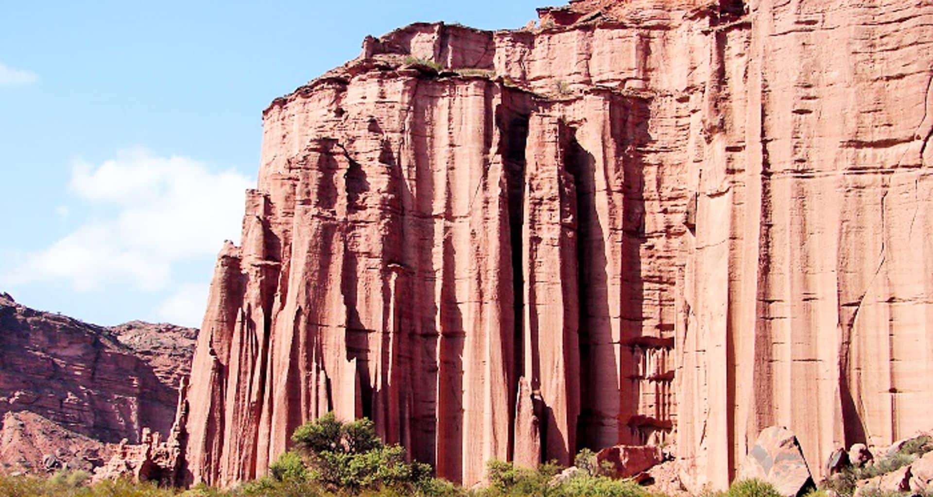 Tall, dramatic red rock cliffs with vertical grooves rise above green shrubs and small trees under a blue sky—scenery reminiscent of Triassic Parks where ancient landscapes shaped the world of early dinosaurs.