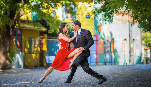 A couple dances the tango outdoors on a cobblestone street near vibrant buildings and green trees, with distant lakes glimmering in the background. The woman wears a red dress and the man a black suit.