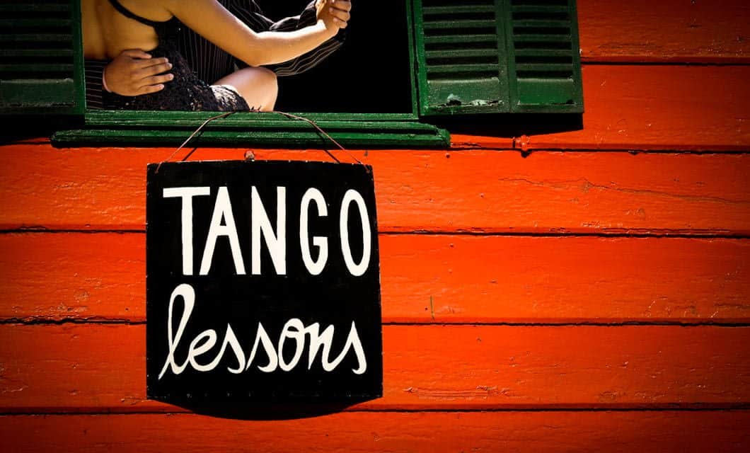 Black sign reading Tango lessons hangs on a bright orange wooden wall below a green-shuttered window, where two people embrace in a tango pose—the spirit of an Argentina Golf Tour captured in this lively street scene.