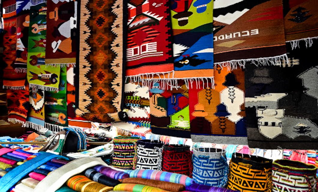 In the heart of the Andes, a vibrant market stall displays colorful woven rugs and textiles with geometric and traditional patterns. Rolled-up fabric and bright blankets are arranged below the hanging rugs.