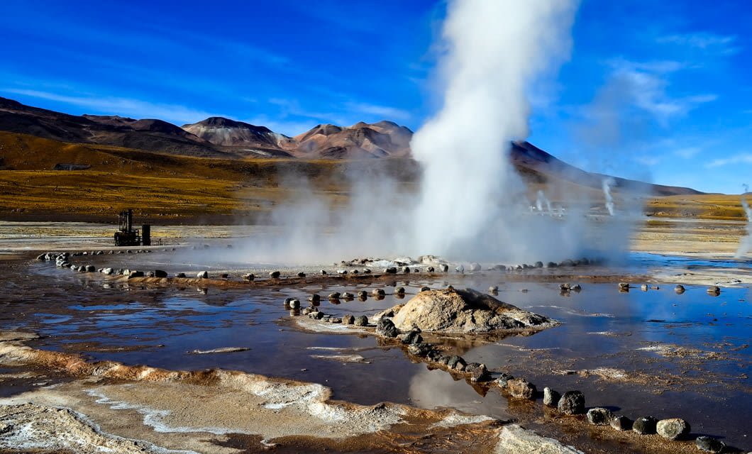 A geothermal geyser shoots steam into the air, surrounded by circular stone markings and shallow pools, set against the majestic Andes mountains and a bright blue sky—nature’s wonder in the heart of the Magic Triangle.