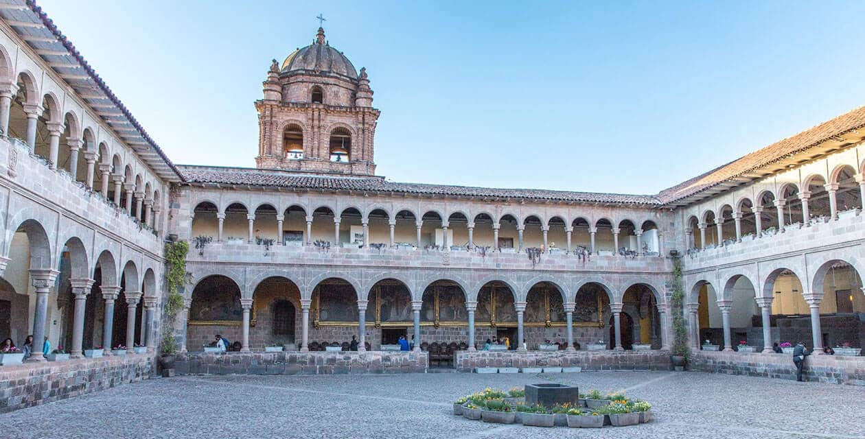 A large stone courtyard with arched walkways lined with columns, surrounding a central fountain and flower beds. A domed bell tower rises above the building under a clear blue sky, evoking the charm of South America's cultural capitals.