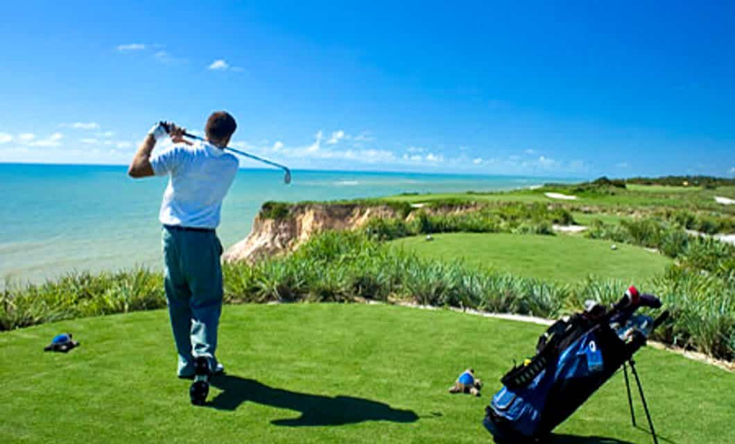 A golfer swings his club on a lush, green course overlooking a scenic coastline with blue ocean and sky—perfect for an unforgettable Argentina Golf Tour. A golf bag stands nearby on the grass.