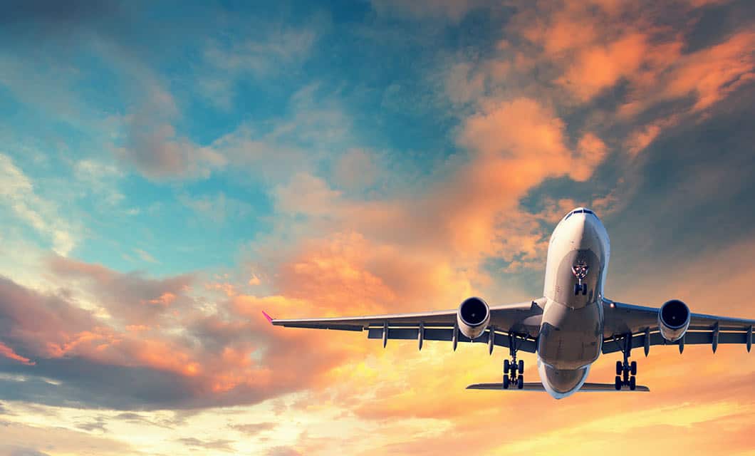 A commercial airplane is flying upwards against a vibrant sky with orange, pink, and blue clouds during sunset—perhaps on its way to a Remote Amazon Tour.
