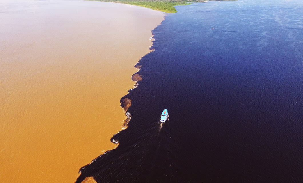 A small boat travels along the distinct meeting point of a muddy brown river and dark blue water on a Remote Amazon Tour, with green land visible in the distance under a clear sky.