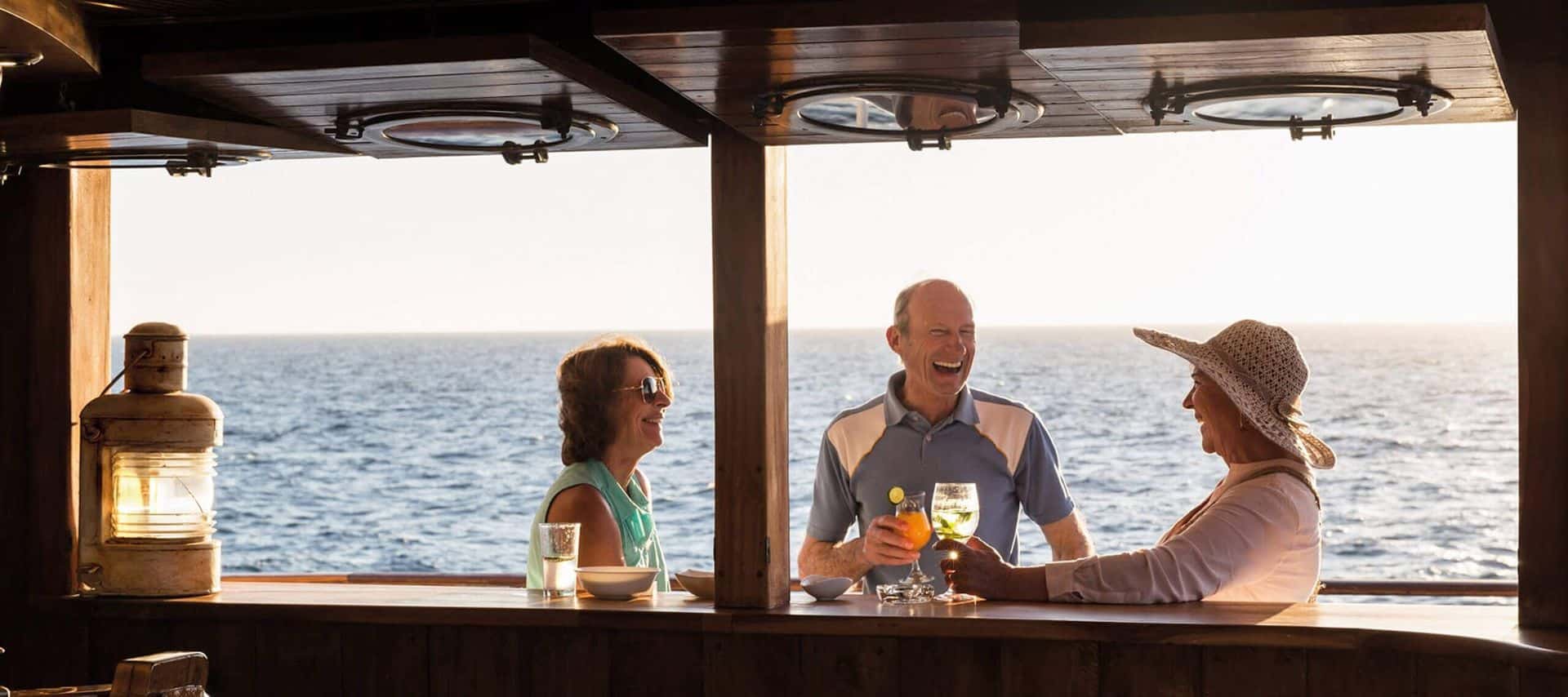 Three older adults smile and chat while enjoying drinks at a shaded bar overlooking the ocean—an ideal scene for relaxing during Ecuador Cruises, with sunlight streaming in and the sea visible in the background.