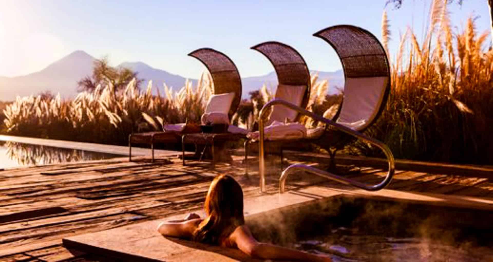 A person relaxes in a steaming outdoor hot tub at sunset at Tierra Atacama Hotel & Spa, with three curved wicker lounge chairs nearby. Tall grasses and a scenic mountain view complete the tranquil setting.