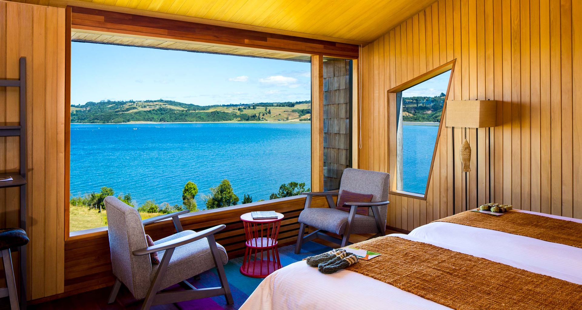 Wood-paneled bedroom at Refúgia Chiloe hotel with large windows overlooking a bright blue lake and green hills, featuring a double bed, two armchairs, small tables, and cozy decor in natural tones—perfect for a relaxing spa retreat.