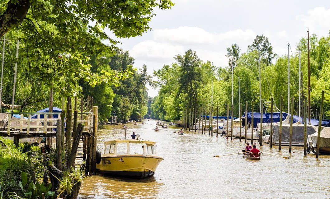 A yellow boat and several small rowboats travel along a wide, tree-lined river in Argentina’s Land of Contrast, with docks, sailboats, and lush green foliage on both sides under a partly cloudy sky.