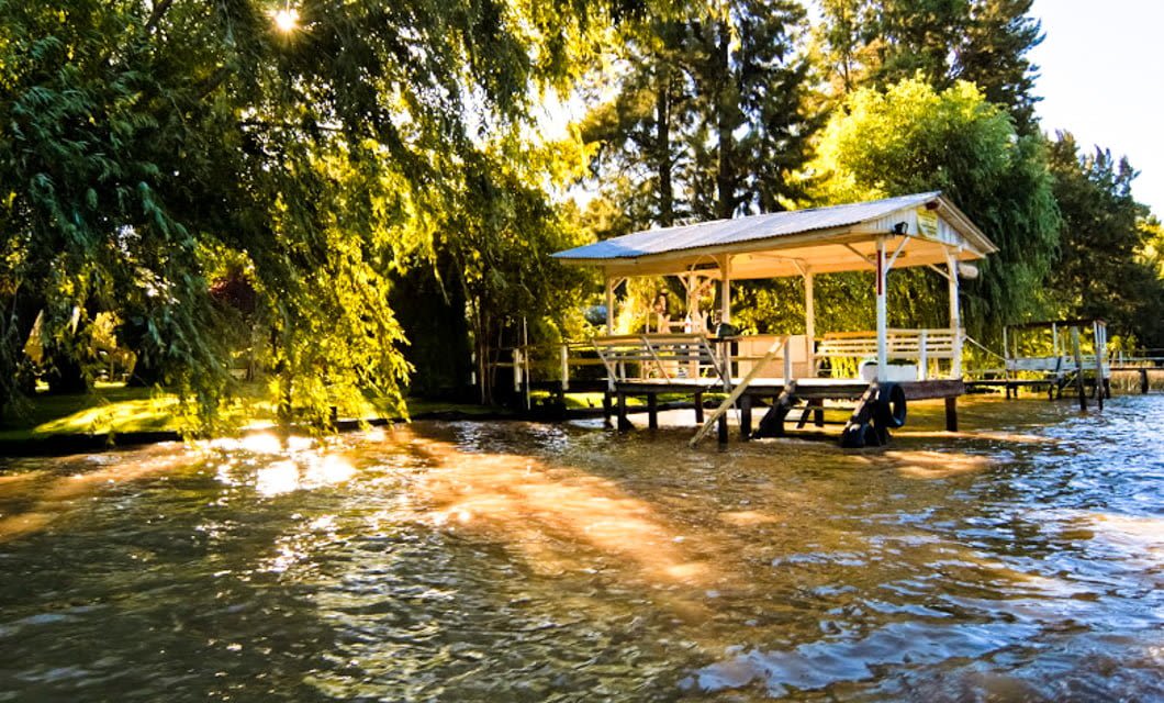 A wooden gazebo with benches stands on stilts over the edge of a sunlit lake, as if awaiting travelers on a Railway Journey, surrounded by lush green trees and reflecting golden sunlight on the rippling water.