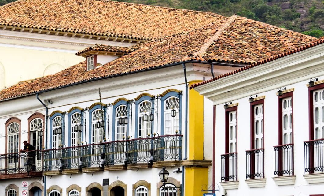 Historic colonial buildings with tiled roofs, colorful blue and yellow window frames, and wrought iron balconies line a street along the historic Route of Gold in Rio de Janeiro, set against a lush green hillside.