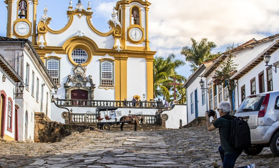A photographer captures a horse-drawn cart in front of a yellow and white baroque church on the Brazil Gold Route, with cobblestone streets lined by colonial-style buildings.