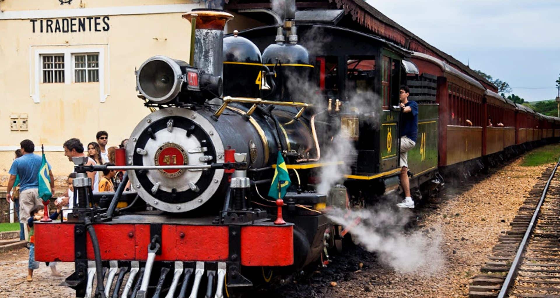 A black and red steam locomotive emits steam at a station labeled Tiradentes, one of Brazil's colonial treasures. Several people stand nearby, some boarding the vintage train while others watch or take photos as it evokes journeys from Rio to Carnival festivities.