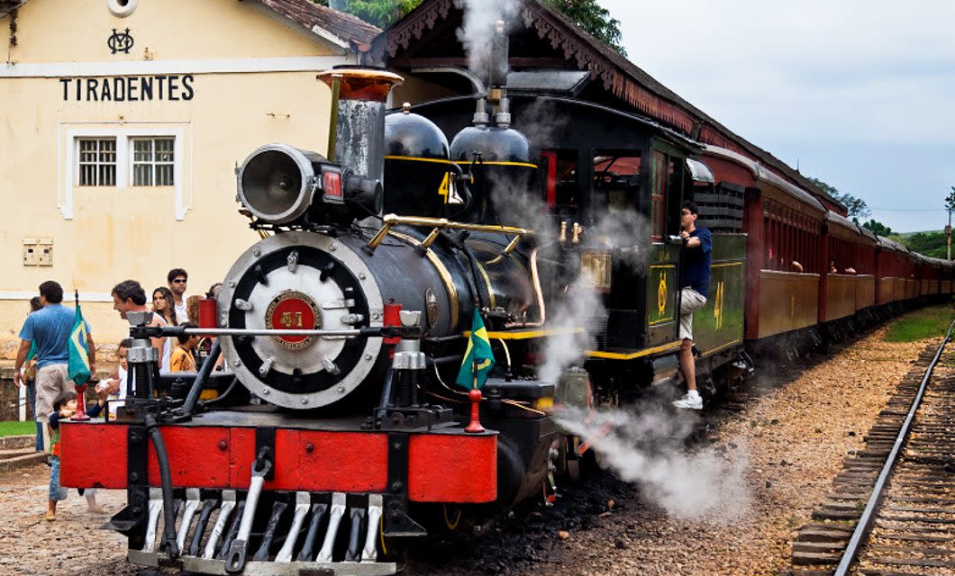 A vintage steam train with a Brazilian flag is stopped at the Tiradentes station. People stand nearby, some boarding or photographing the train, as steam rises around the locomotive—exploring one of Brazil’s Colonial Treasures.