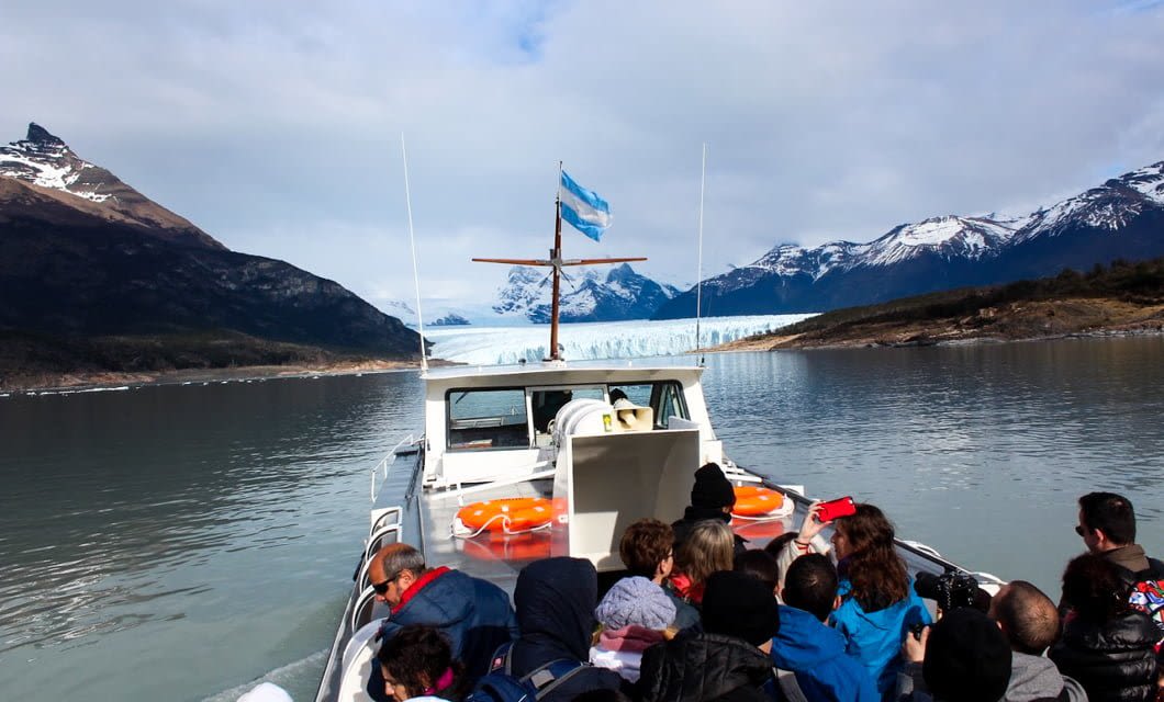 A group of people ride on a boat toward a massive glacier in Patagonia, surrounded by mountains under a partly cloudy sky. The boat, part of Skorpios Cruises, has an Argentine flag and bright orange life rings on its deck.