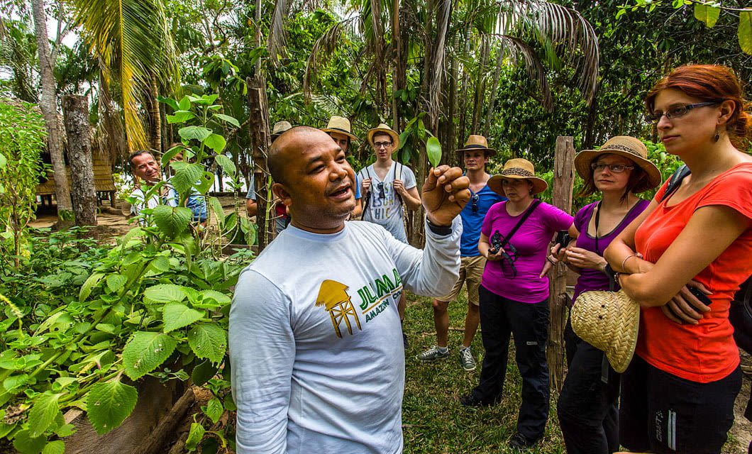 A man in a white shirt explains a plant to a group of attentive people on a Brazil Tour in Salvador Bahia’s lush, tropical garden; some listen, others take notes or photos, and most wear hats and casual clothes.