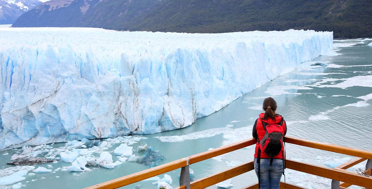 A person wearing a red jacket and backpack stands on a wooden viewing platform in Patagonia, gazing at a vast blue glacier that stretches across a lake toward forested mountains—one of the must-see Patagonia essentials.