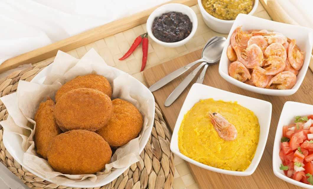 A tray with acarajé (fried patties), bowls of shrimp, yellow mush, chopped tomatoes, and two sauces—all inspired by a tour through Brazil's Cerrado. Two red chili peppers and utensils are placed beside the dishes.
