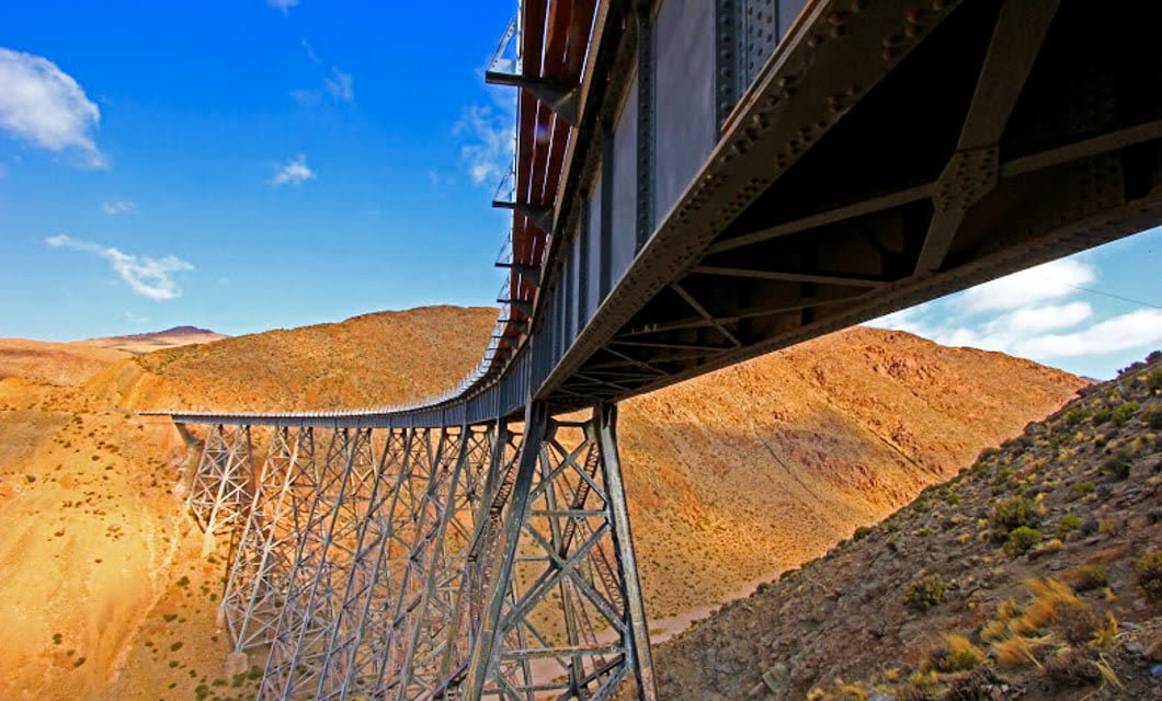 A large metal railway bridge curves over a dry, mountainous landscape under a bright blue sky with scattered clouds, reminiscent of the iconic Train to the Clouds railway journey.