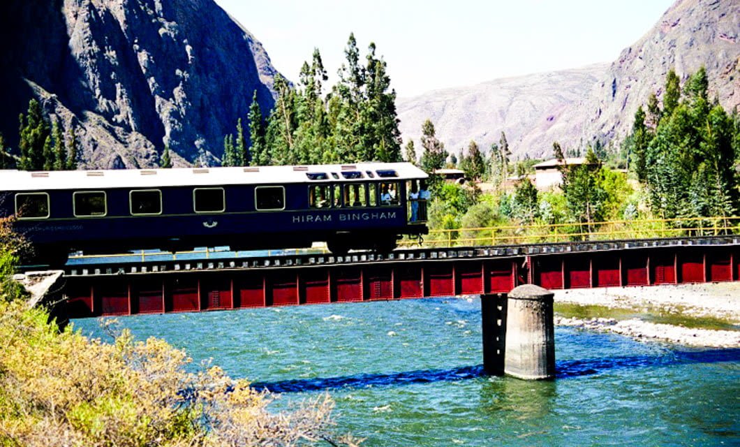 A blue train car labeled Hiram Bingham crosses a red bridge over a river, surrounded by green trees and tall mountains under a clear sky—an iconic tour en route to Machu Picchu.