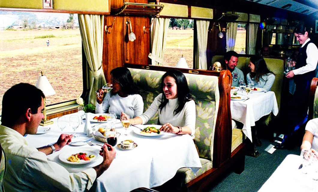 People dining in a luxurious train dining car during a Peru Bolivia Rail Tour, enjoying meals and drinks at tables with white tablecloths as a server attends to guests. Large windows reveal a scenic landscape outside.