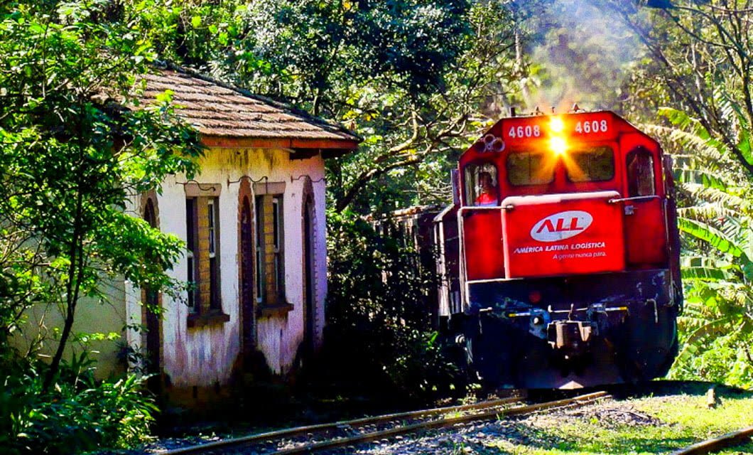 A red locomotive on a Brazil Train Tour passes by an old, weathered house surrounded by dense green vegetation and trees, traveling along a narrow railway track in bright daylight.