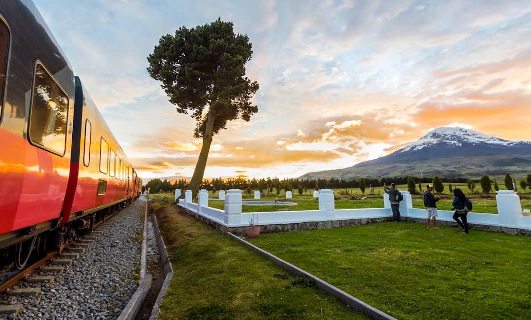 A red and black Tren Crucero train travels past a green lawn with people taking photos, white stone markers, a leaning tree, and a snow-capped mountain in the Avenue of the Volcanoes under a colorful sunset sky.