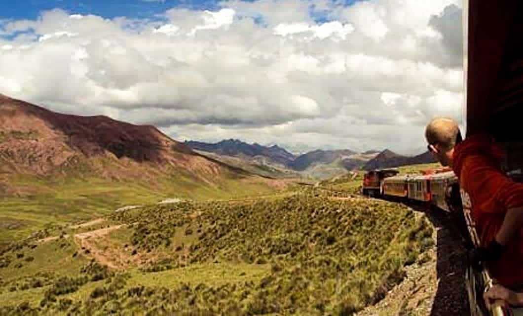 A person leans out of a moving train on a scenic Train Tour through Bolivia or Peru, admiring green mountain scenery under a cloudy sky as the train curves along tracks through a vast landscape with distant peaks.