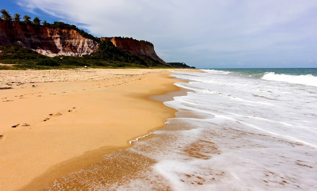 Golden sandy beach with gentle waves washing ashore, bordered by lush vegetation and tall, reddish cliffs under a partly cloudy sky. Experience this serene and natural scene as part of your Argentina Golf Tour adventure.