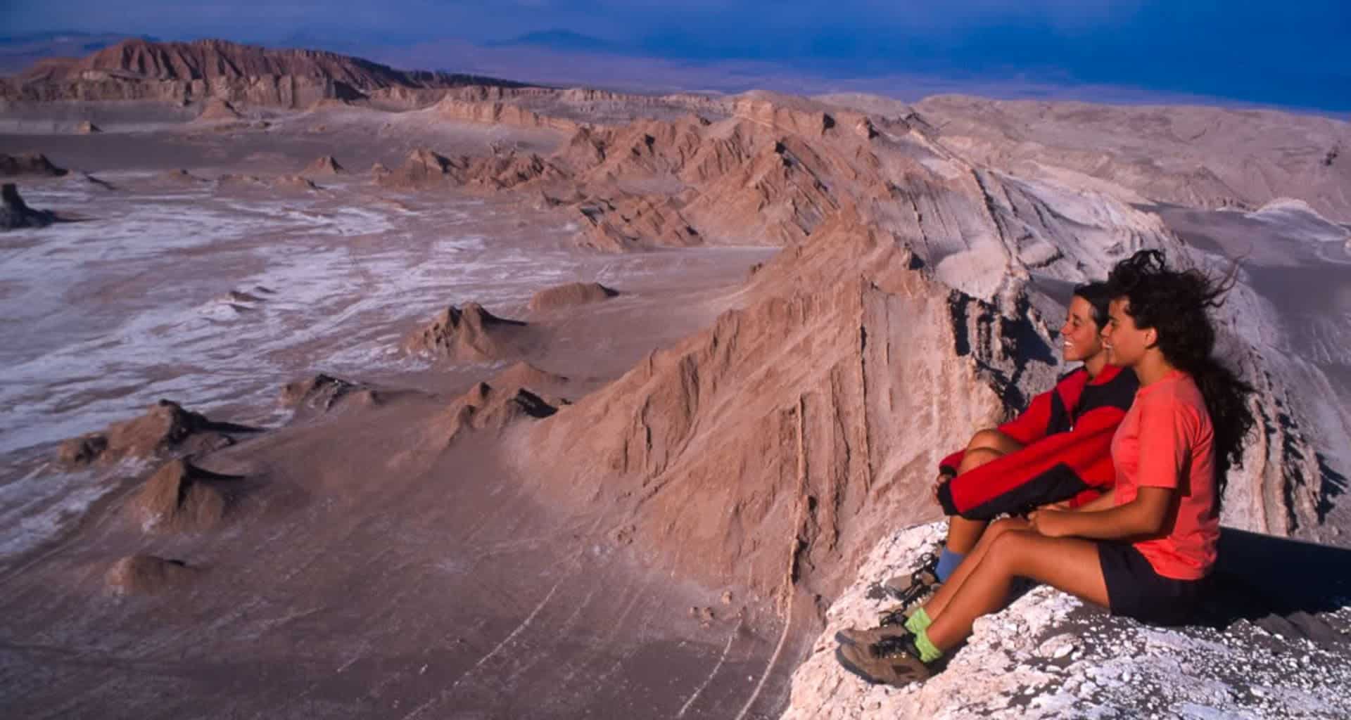 Two people sit on the edge of a rocky cliff, overlooking the vast, arid landscape of the Magic Triangle of the Andes, with rugged hills and sandy terrain stretching beneath a clear blue sky.