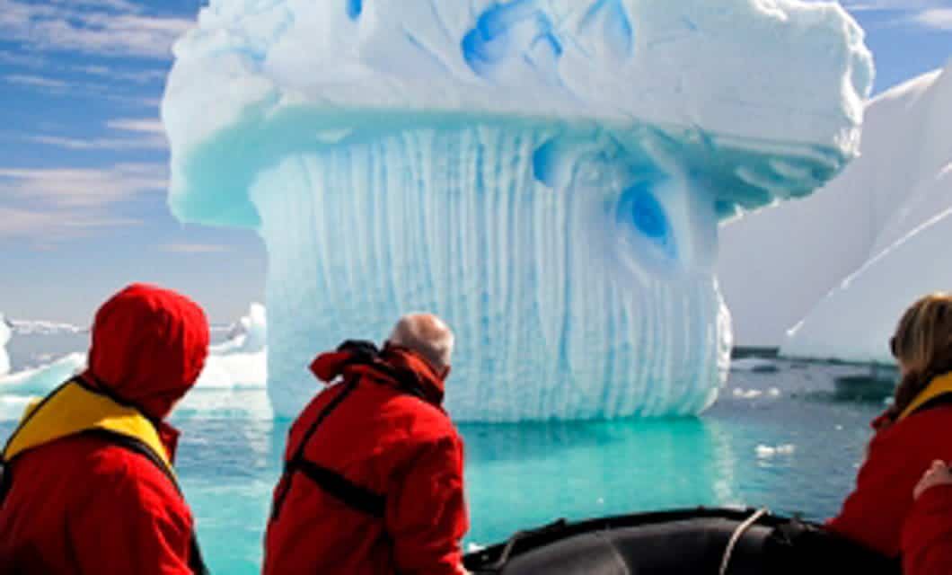 Three people in red jackets on a small boat admire a large, uniquely shaped iceberg with vertical grooves, drifting in turquoise water under a partly cloudy sky during an Antarctica cruise from Ushuaia aboard the Plancius.