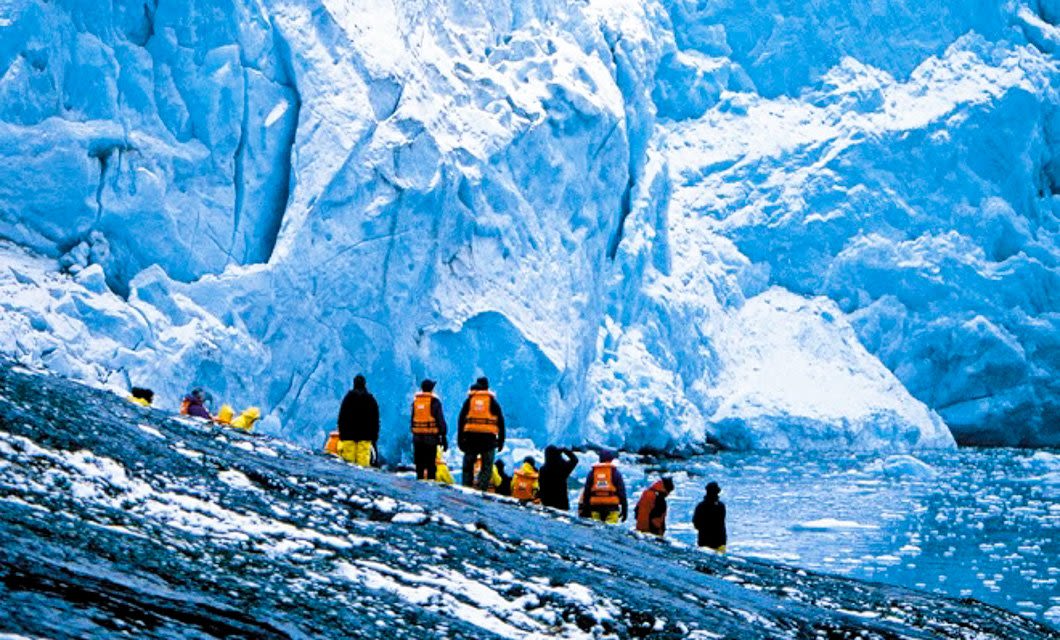 A group of people wearing orange jackets stand on a rocky, icy shore in the Fuegian Fjords of Patagonia, in front of towering blue-white glaciers beside a cold body of water.