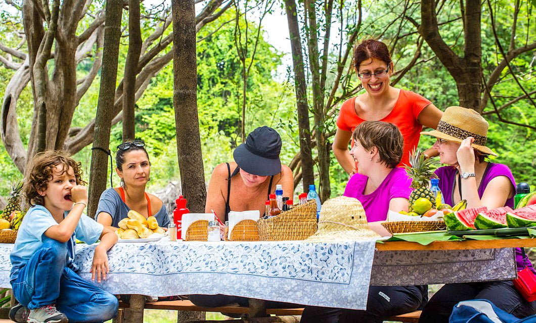 A group of people gather around an outdoor picnic table covered with fruit, drinks, and baskets in the shade—enjoying laughter and conversation, just like a relaxed afternoon in Salvador Bahia. A young child sits on the table’s edge eating a snack.