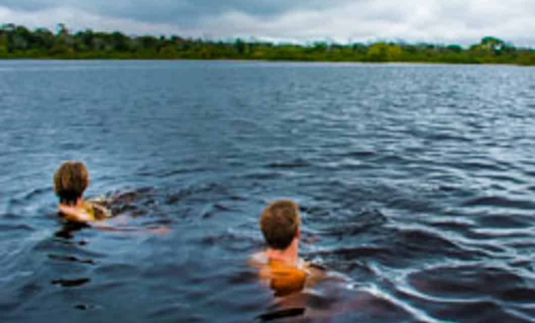 Two people are swimming in a large, dark blue lake under a cloudy sky, with forested land visible in the background—an experience reminiscent of Amazon expeditions offered by Tahuayo Lodge on many Peru tours.