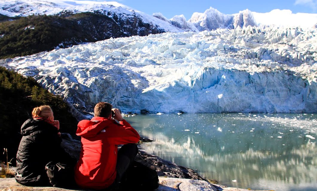 Two people in winter jackets sit on rocks, gazing at a large glacier and snow-capped mountains reflected in a calm lake under a clear blue sky—an awe-inspiring scene from Tierra del Fuego.