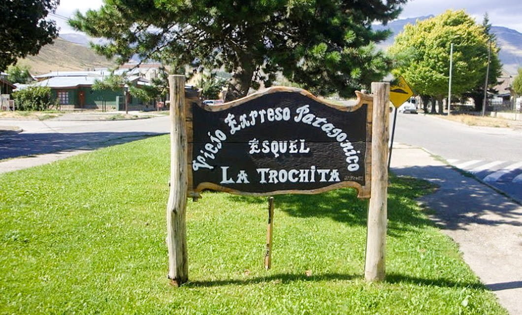Wooden sign on a grassy area reads “Viejo Expreso Patagónico, Esquel, La Trochita,” marking the historic Patagonia Train tour route, with a street, trees, buildings, and distant mountains in the background.